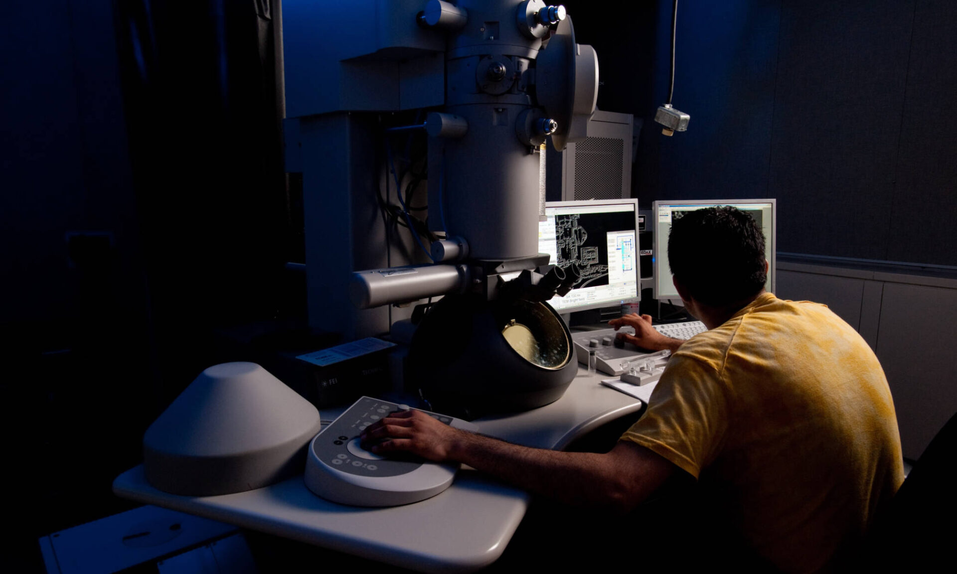 A person in a yellow shirt seen from behind works with a transmission electron microscope in a darkened room.